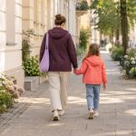 Eine erwachsene Person und ein Kind gehen Hand in Hand auf einem sonnigen Gehweg, von hinten betrachtet