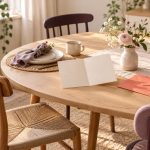 Unbeschriftete Einladung und korallfarbene Umschläge auf einem einladend gedeckten Tisch mit unterschiedlichen Stühlen.