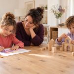 Zwei Kinder zeichnen und bauen an einem Holztisch. Eine erwachsene Person begleitet das zeichnende Kind aufmerksam.